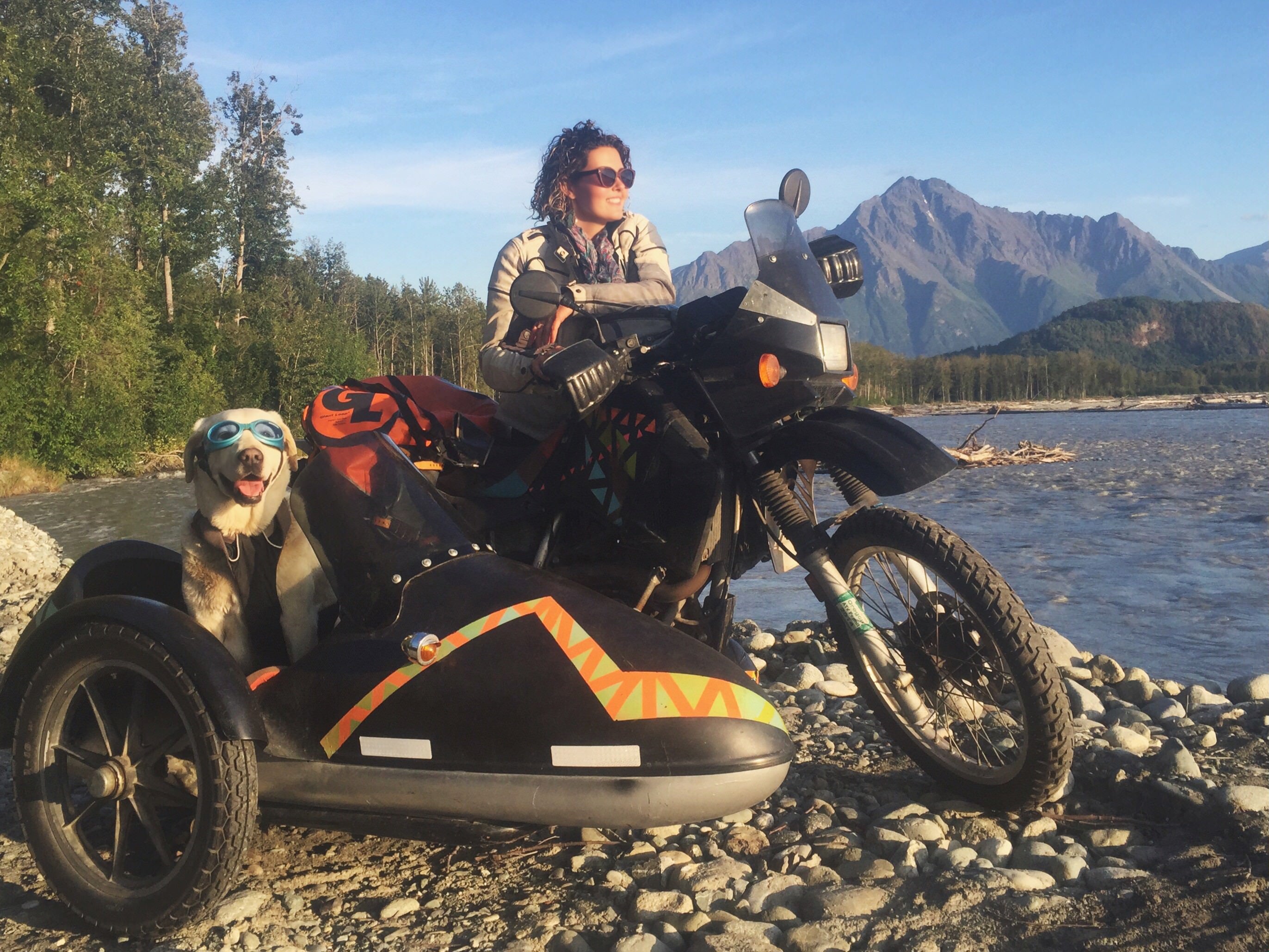 Person named Mallory Paige riding a motorcycle with a sidecar featuring a dog, set against a scenic mountain and water backdrop in Alaska