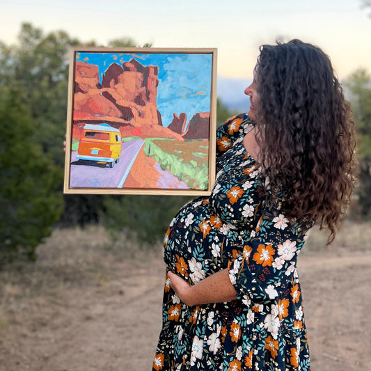 A woman named Mallory Paige holding a framed painting of a desert scene with a VW campervan.