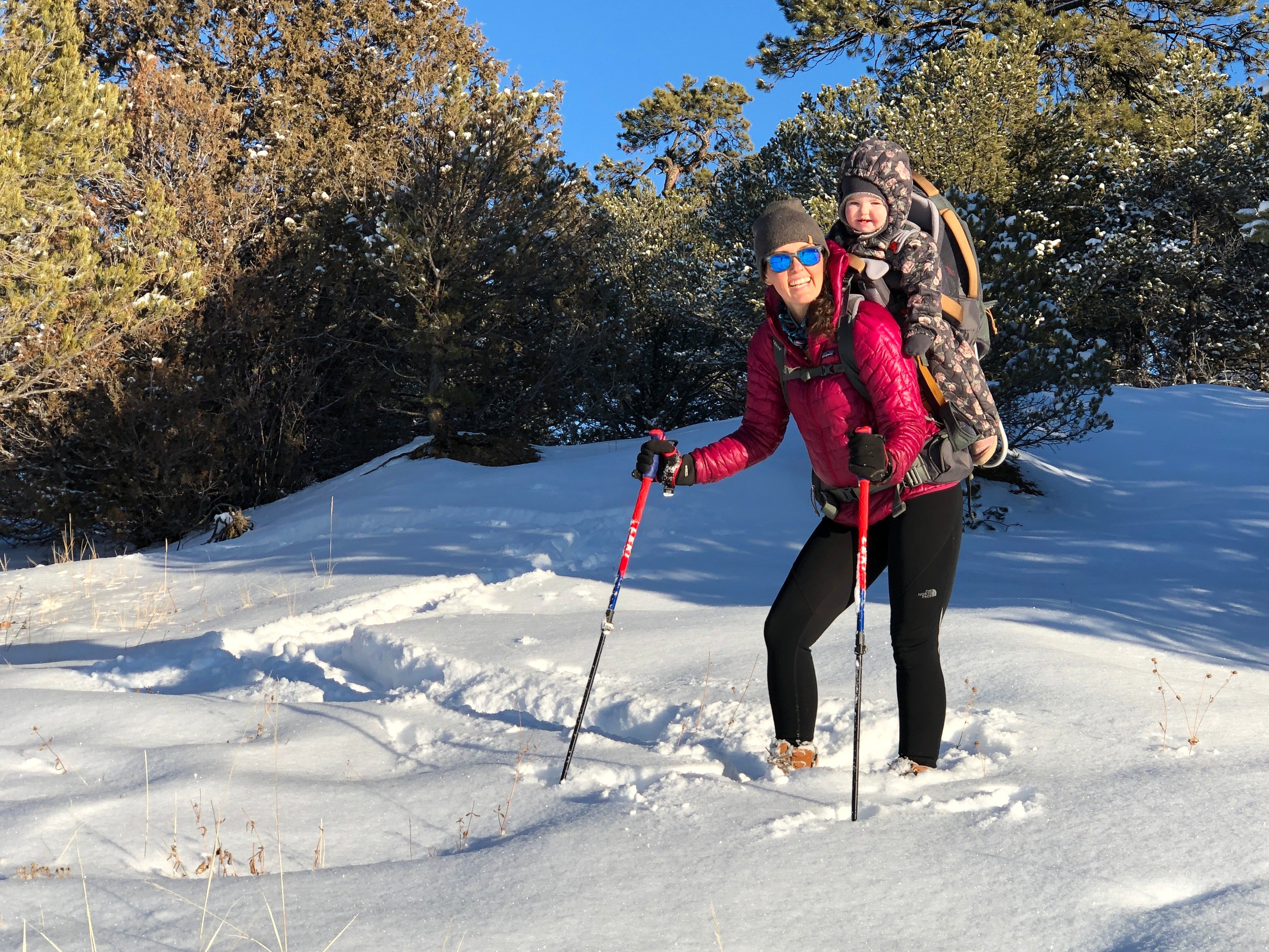 A woman named Mallory Paige hiking in the snow with a child on their back, surrounded by trees.