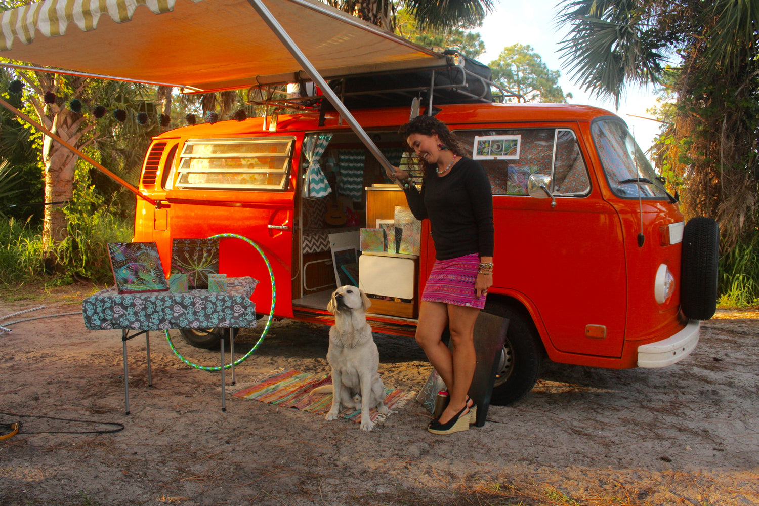 An woman named Mallory Paige standing next to an orange van with an extended awning, surrounded by outdoor furniture and a dog.