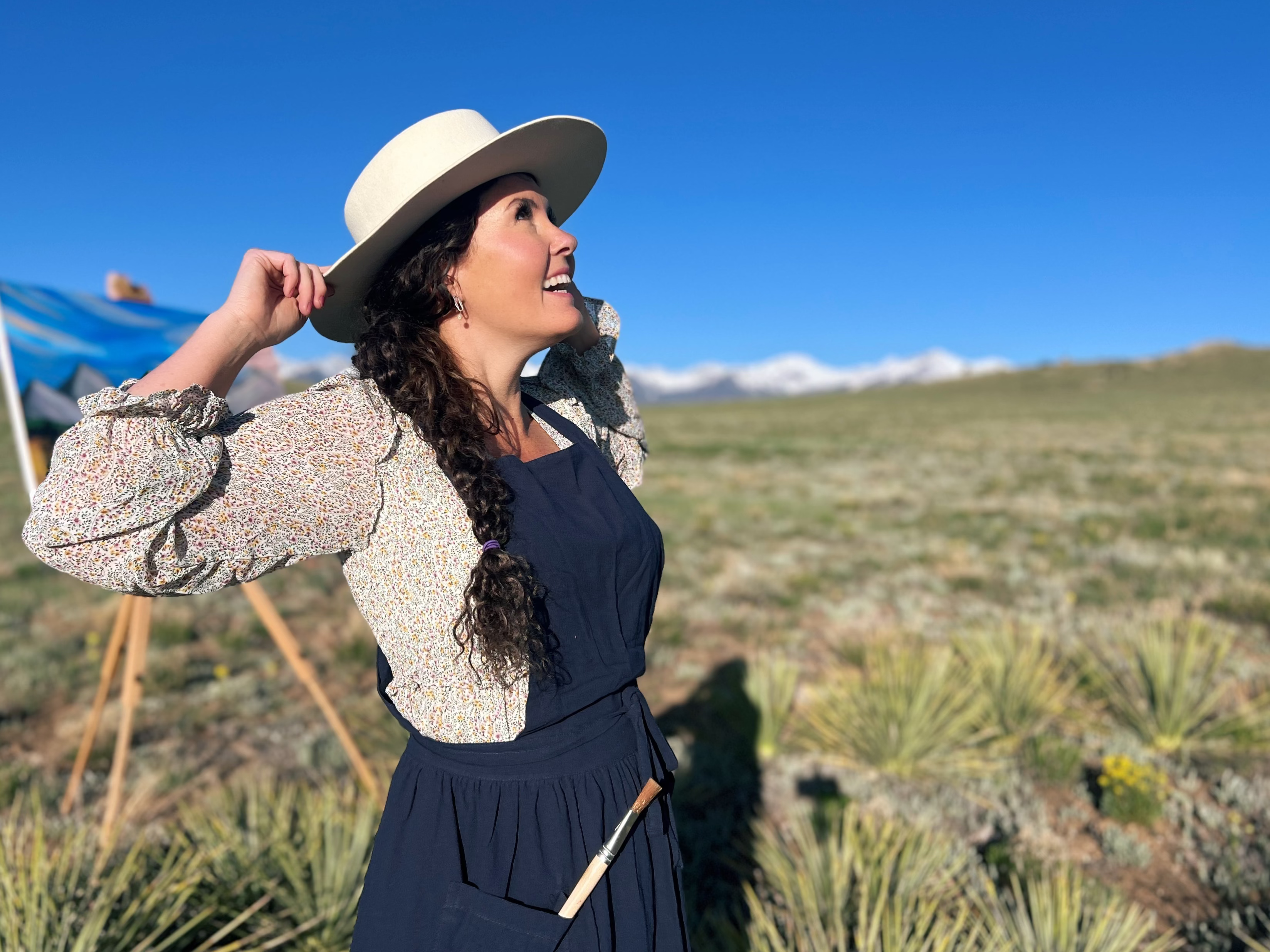 Painter Mallory Paige  in a hat and dress standing in a field with mountains in the background and art on an easel.